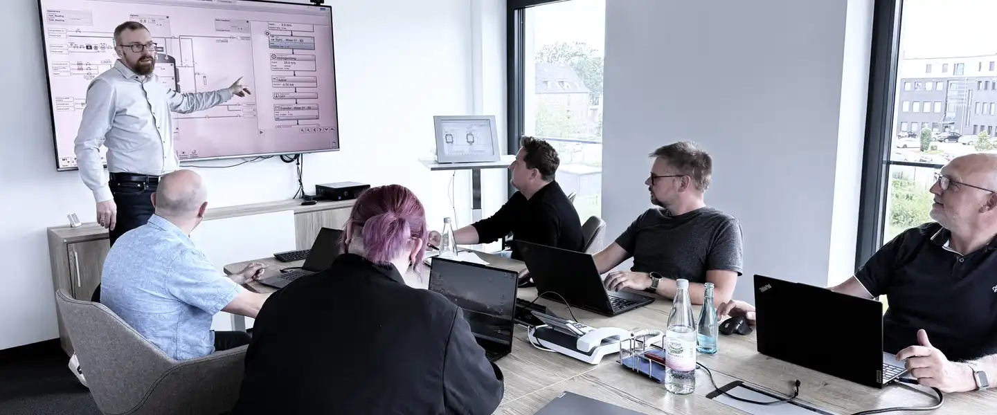 Eine Gruppe von Personen in einem Besprechungsraum, einige heben die Hand, während jemand an einem Bildschirm präsentiert.