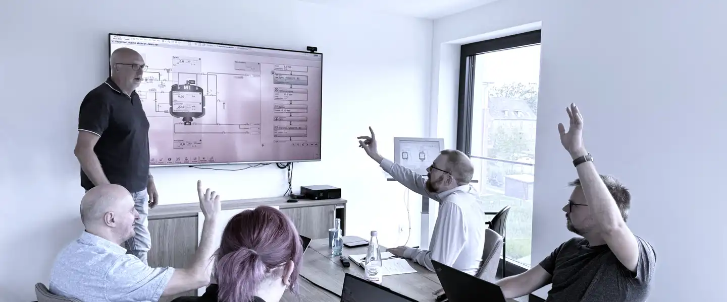 Eine Gruppe von Personen in einem Besprechungsraum, einige heben die Hand, während jemand an einem Bildschirm präsentiert.